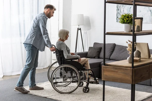 Ganzkörperansicht eines Mannes, der behinderte Mutter im Rollstuhl trägt — Stockfoto