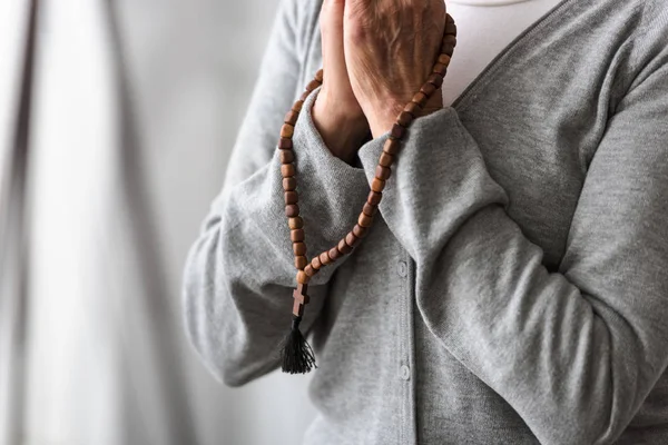 Partial view of senior woman praying with wooden rosary — Stock Photo