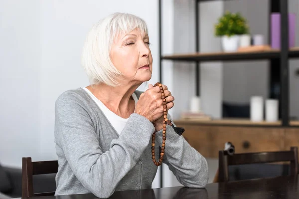 Seniorin hält hölzernen Rosenkranz und betet mit geschlossenen Augen — Stockfoto