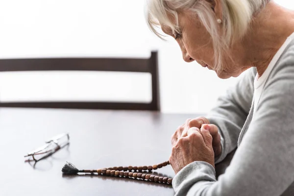 Mujer mayor sosteniendo rosario de madera y orando con los ojos cerrados - foto de stock