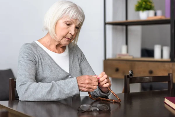 Nachdenkliche Seniorin sitzt am Tisch und hält Rosenkranz in der Hand — Stockfoto