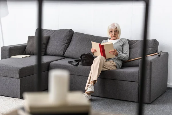 Mulher sênior em óculos sentado no sofá e livro de leitura na sala de estar — Fotografia de Stock