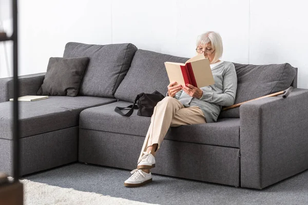 Seniorin mit Brille sitzt auf Sofa und liest Buch im Wohnzimmer — Stockfoto