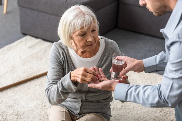 Vista parcial del hombre dando a la madre mayor vaso de agua y medicina - foto de stock