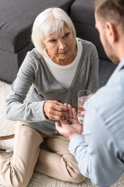 Vista parcial del hombre dando a la madre mayor vaso de agua y medicina - foto de stock