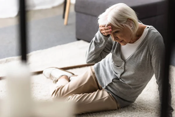 Пожилая женщина с мигренью сидит на ковре и трогает лоб рукой — стоковое фото