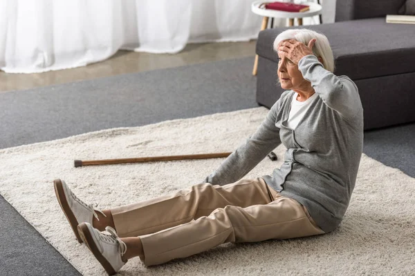 Donna anziana con emicrania seduta su tappeto e toccare la fronte con mano — Foto stock