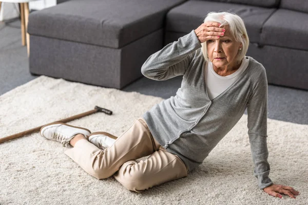 Пожилая женщина с мигренью сидит на ковре и трогает лоб рукой — стоковое фото
