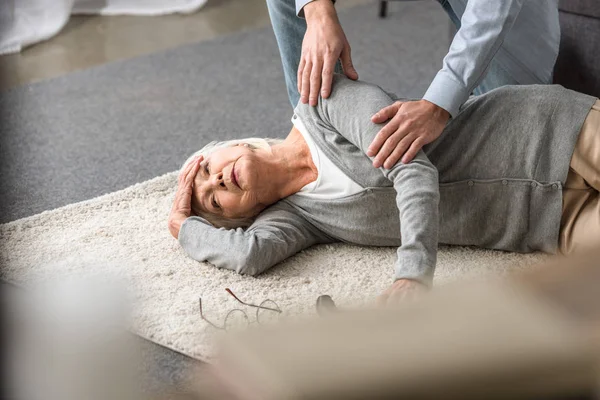 Visão cortada do homem ajudando a mãe sênior com ataque cardíaco caído no tapete — Fotografia de Stock