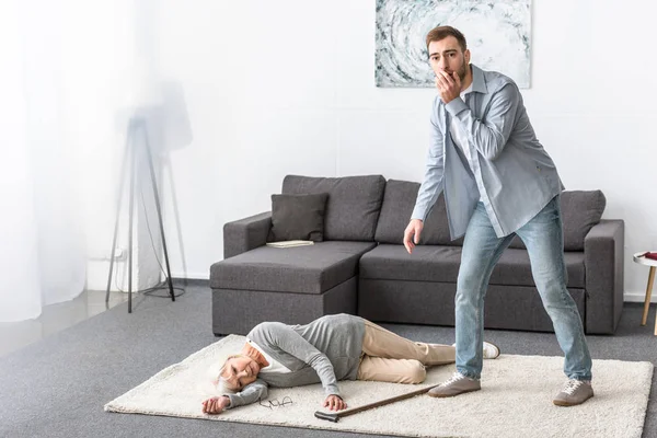 Full length view of worried man standing near senior woman lying on carpet — Stock Photo