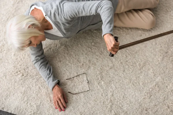 Overhead view of sick senior woman with walking stick lying on carpet — Stock Photo