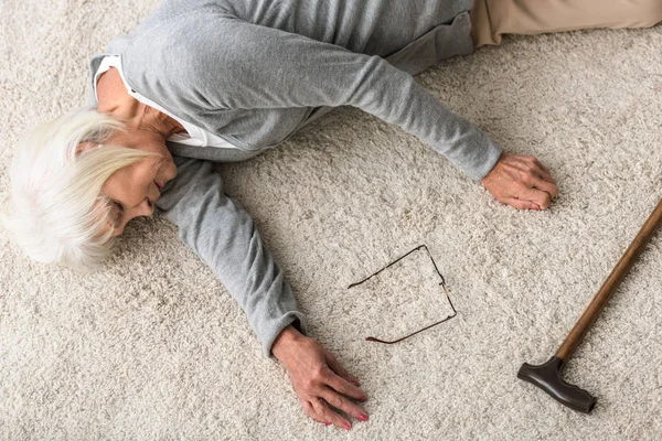 Overhead view of sick senior woman with walking stick lying on carpet — Stock Photo