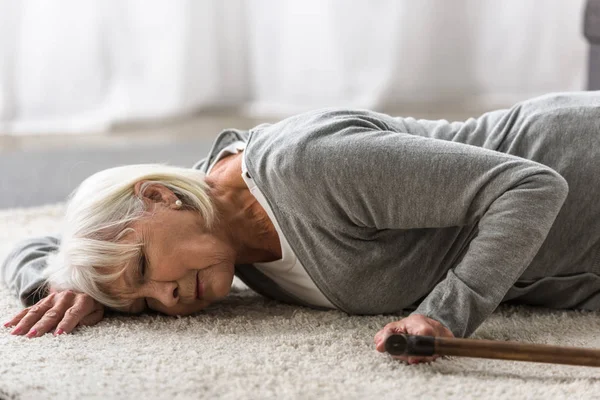 Ill senior woman with cane lying on carpet with closed eyes — Stock Photo