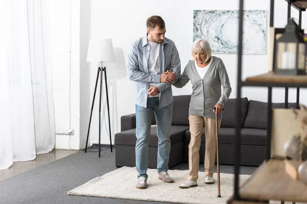 Vista completa del hombre ayudando a la madre mayor con bastón en casa - foto de stock