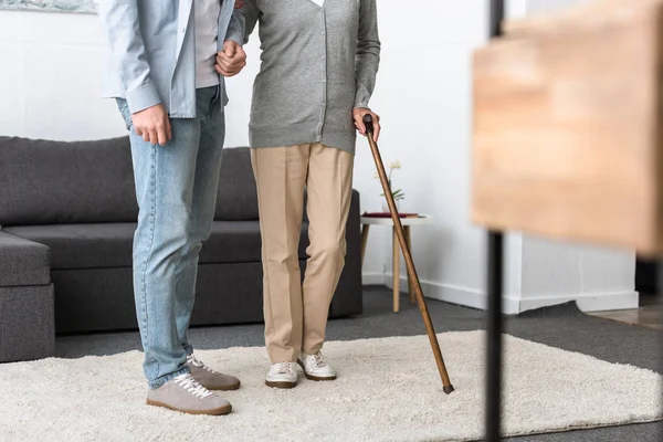 Vista recortada del hombre ayudando a la madre mayor con bastón en casa - foto de stock