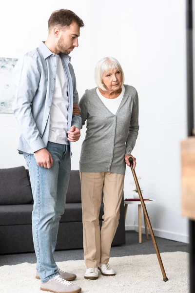В полный рост мужчина помогает пожилой матери с тростью дома — стоковое фото