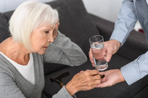 Ausgeschnittene Ansicht eines Mannes, der der Seniorin ein Glas Wasser und Medizin schenkt — Stockfoto