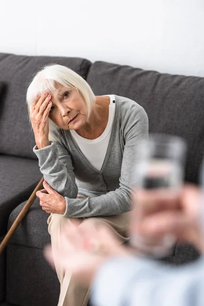 Messa a fuoco selettiva dell'uomo che tiene il bicchiere d'acqua e la donna anziana seduta sul divano — Foto stock