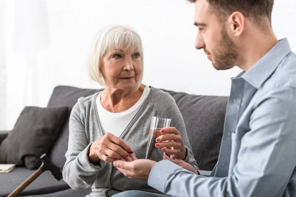 Cropped view of man giving to senior mother glass of water and medicine — Stock Photo