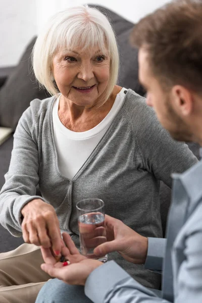 Обрезанный вид человека, дающего старшей матери стакан воды и таблетки — стоковое фото