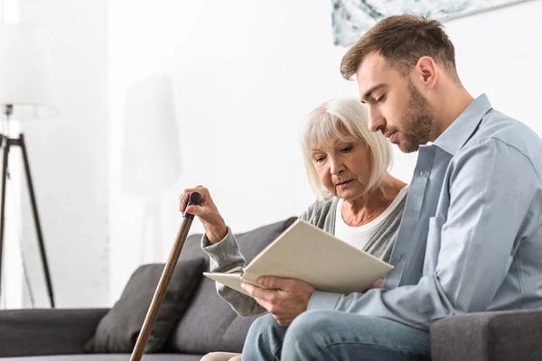 Hombre sentado en el sofá y libro de lectura con la madre mayor - foto de stock