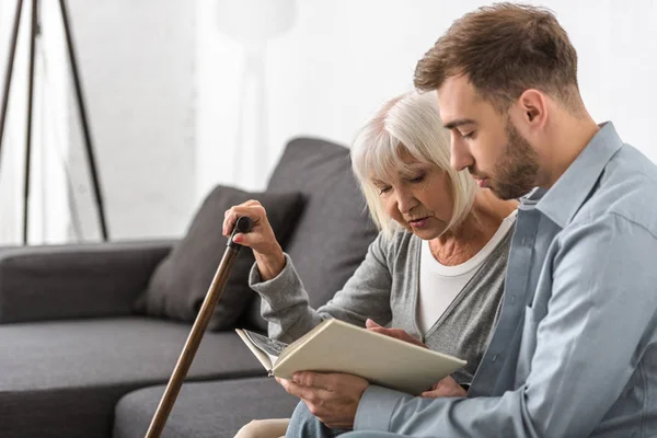 Mann sitzt mit älterer Mutter auf Sofa und liest Buch — Stockfoto