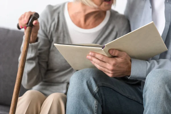 Cropped view of man sitting on sofa and reading book with mother — Stock Photo