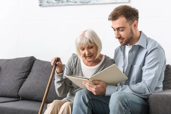 Uomo seduto sul divano e libro di lettura con la madre anziana — Foto stock