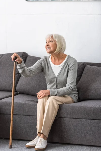 Femme âgée souriante avec canne assis sur le canapé et regardant loin dans le salon — Photo de stock