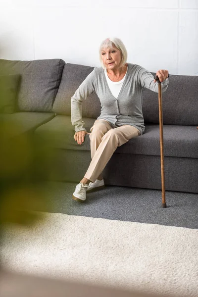 Senior woman with walking stick sitting on sofa in living room — Stock Photo