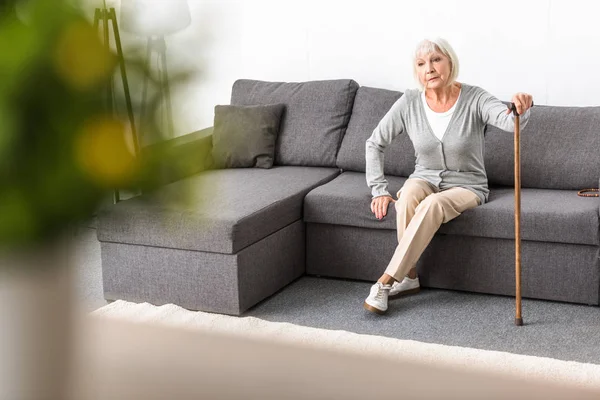 Femme âgée avec bâton de marche assis sur le canapé dans le salon — Photo de stock