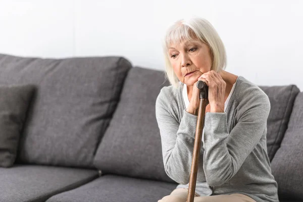 Pensive senior woman with cane sitting on sofa and looking down — Stock Photo