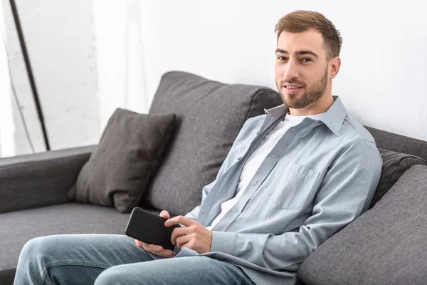 Бородатий чоловік сидить на дивані і тримає смартфон у вітальні — стокове фото