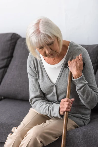 Pensive senior woman with wooden cane sitting on sofa in living room — Stock Photo