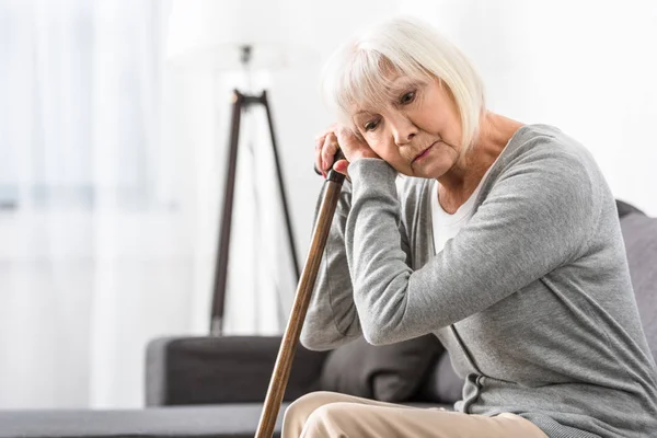 Pensive senior woman with wooden cane sitting on sofa in living room — Stock Photo