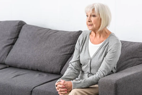 Nachdenkliche Seniorin mit grauen Haaren sitzt auf Sofa im Wohnzimmer — Stock Photo