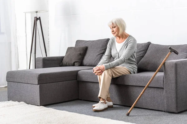 Pensive senior woman with wooden cane sitting on sofa in living room — Stock Photo