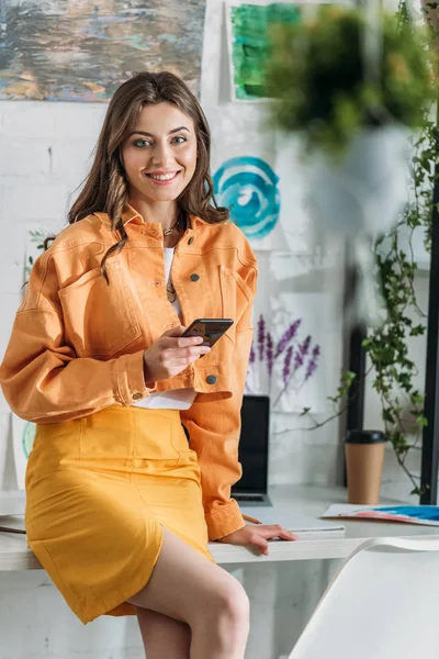 Selective focus of pretty fashionable woman looking at camera while smiling and holding smartphone — Stock Photo