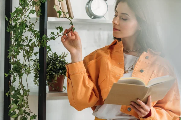 Selective focus of pensive young woman holding book and touching plant while standing bu rack — Stock Photo