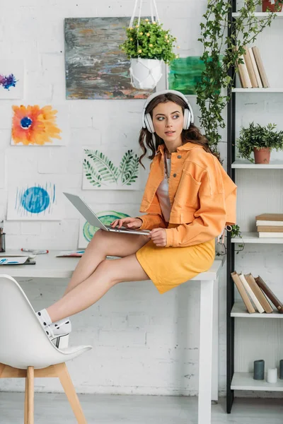 Trendy young woman in headphones using laptop while sittig on desk at home — Stock Photo