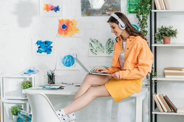 Mujer sonriente de moda en los auriculares usando el ordenador portátil mientras está sentado en el escritorio - foto de stock