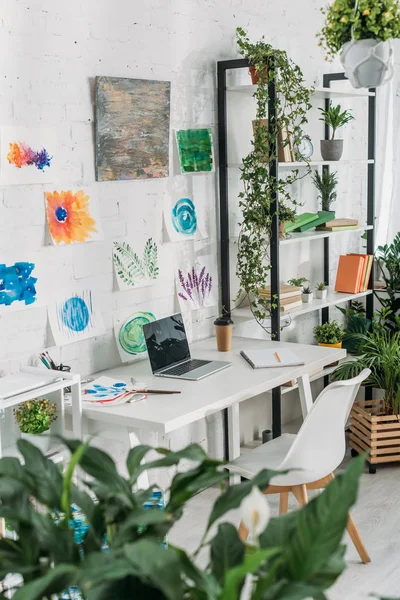 Enfoque selectivo de enfoque selectivo de habitación espaciosa con ordenador portátil en la mesa, pinturas en la pared y estante con plantas en maceta — Stock Photo