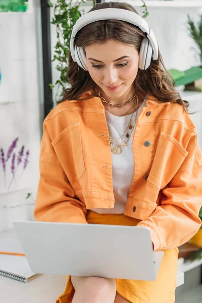 Симпатичная улыбчивая девушка слушает музыку в наушниках и пользуется ноутбуком дома — стоковое фото