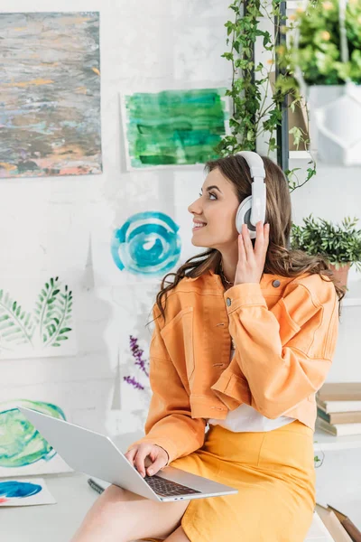 Femme heureuse regardant des peintures sur le mur tout en écoutant de la musique dans les écouteurs et en utilisant un ordinateur portable — Photo de stock
