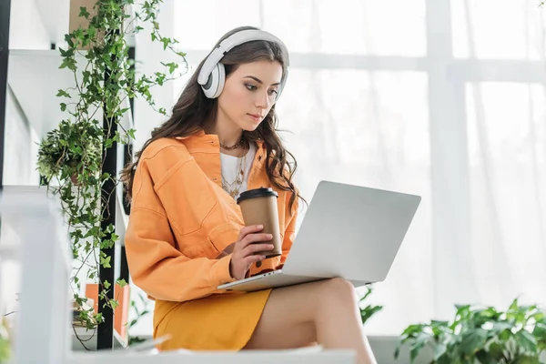 Aufmerksame junge Frau hält Kaffeebecher in der Hand, bedient Laptop und hört Musik über Kopfhörer — Stockfoto