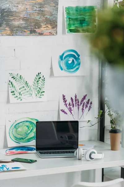 Selektiver Fokus des Laptops mit leerem Bildschirm, Kopfhörer und Pappbecher auf Tischplatte in der Nähe von Gemälden — Stockfoto