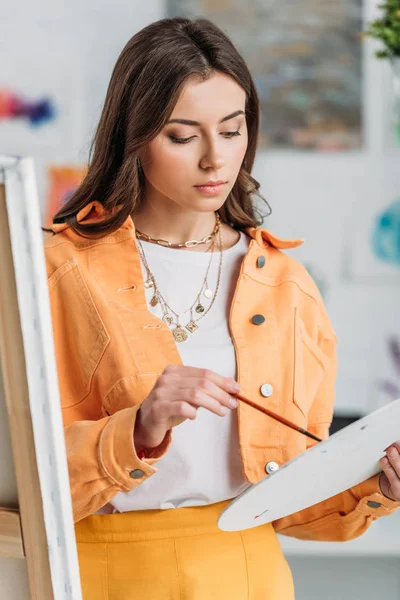 Attractive pensive artist holding palette and paintbrush while standing near easel — Stock Photo