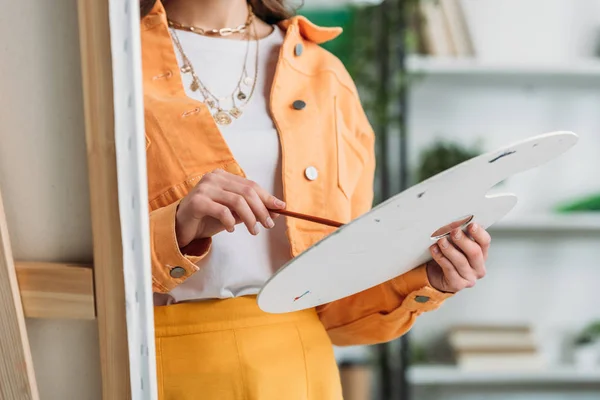 Partial view of young artist holding palette and paintbrush while standing near easel — Stock Photo