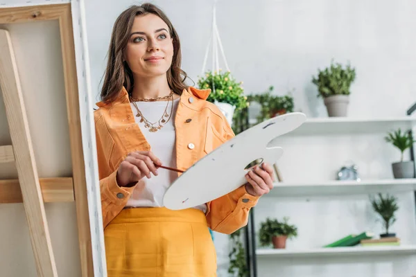 Smiling young artist looking away while standing near easel with palette — Stock Photo