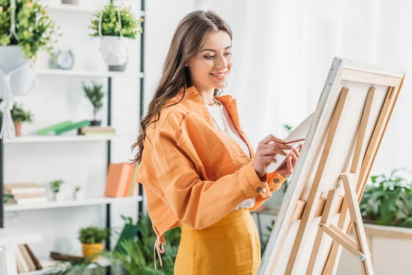 Jovem artista atraente sorrindo enquanto desenho sobre tela com pincel — Fotografia de Stock